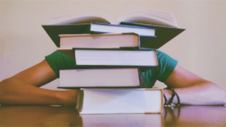 student sitting at a table, obscured by a stack of books.